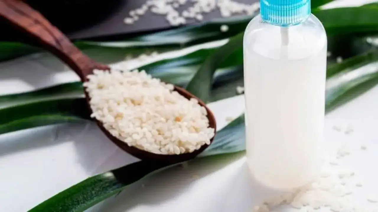 Cuchara de madera con arroz y botella con agua de arroz para la cara, rodeado de hojas verdes.