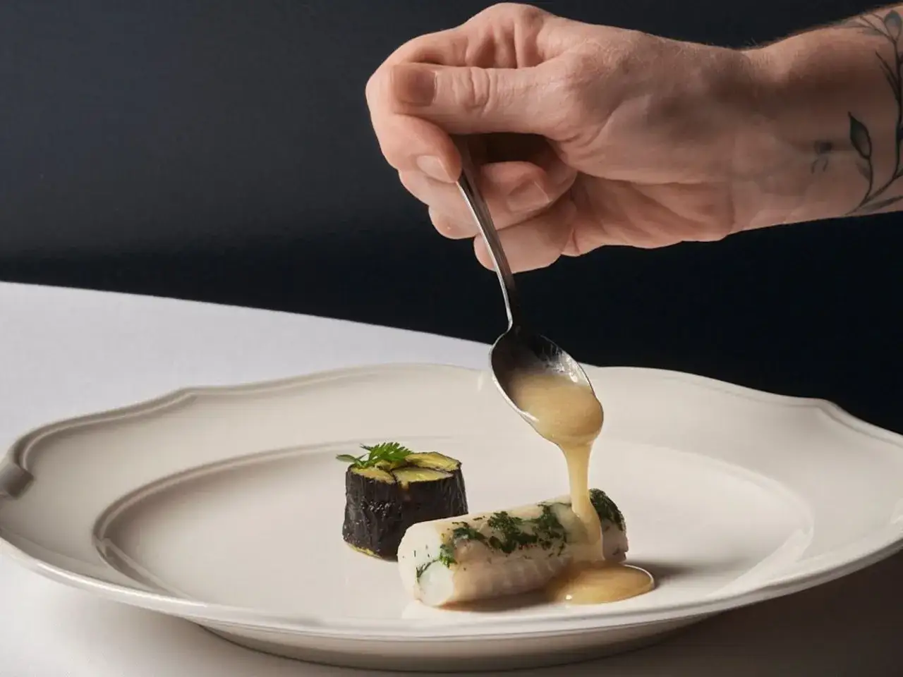 Un cuoco versa una salsa dorata su un filetto di sogliola alla mugnaia, accompagnato da un rotolo di melanzane.