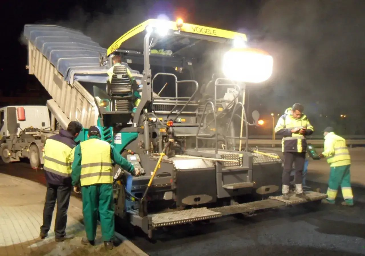 Trabajadores operan una pavimentadora V&ouml;gele por la noche, descargando material caliente. El proceso de pavimentaci&oacute;n con slurry es esencial para la construcci&oacute;n de carreteras.