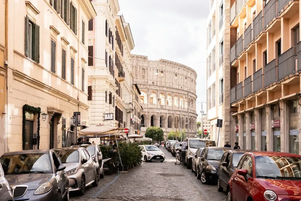 Straßenansicht in Rom mit dem Kolosseum im Hintergrund. Autos parken am Straßenrand, ein Bar-Schild ist sichtbar. Ein typischer Tag in Italien.