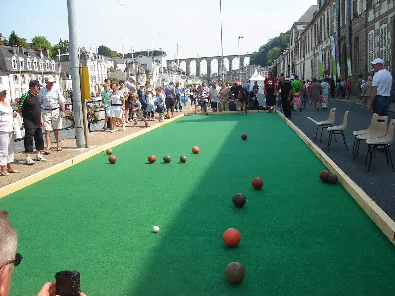 Des joueurs s'affrontent sur un terrain de pétanque, une **règle boule bretonne** est visible. Des spectateurs observent la partie.