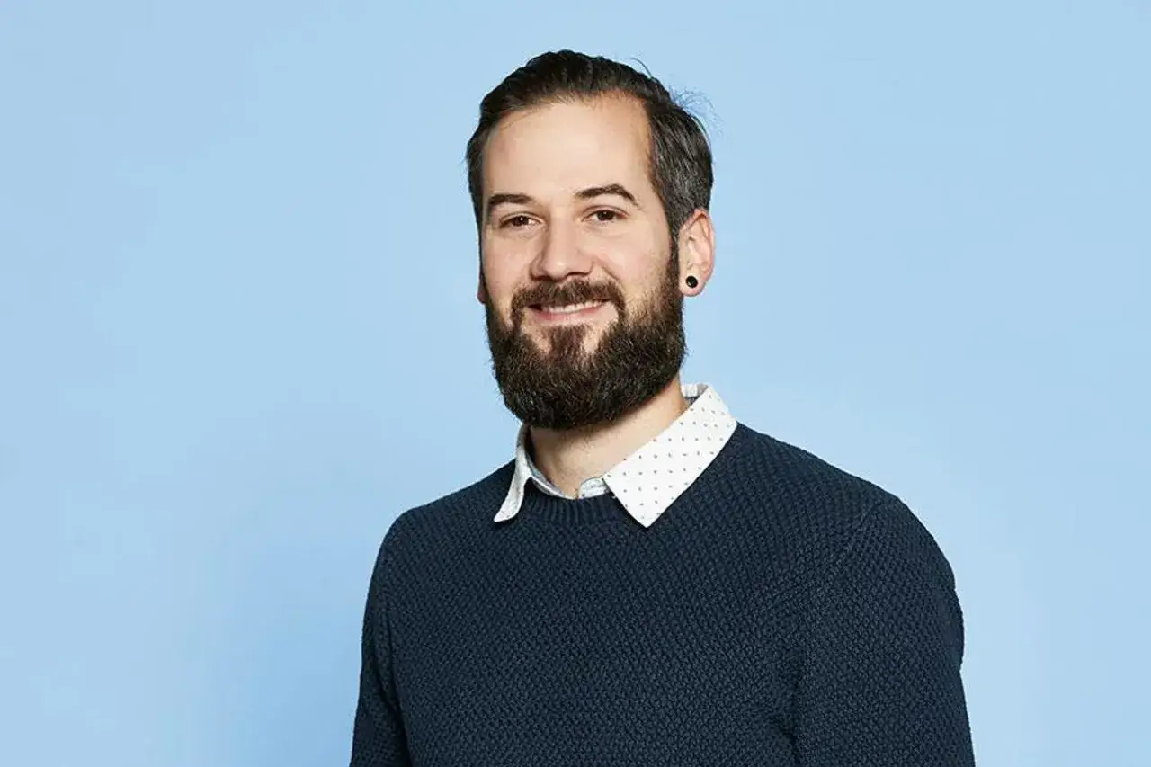 Ein Mann mit Bart und Ohrstecker, der einen blauen Pullover &uuml;ber einem Hemd tr&auml;gt. Er k&ouml;nnte ein sinnfluencer deutschland sein, der &uuml;ber positive Themen spricht.