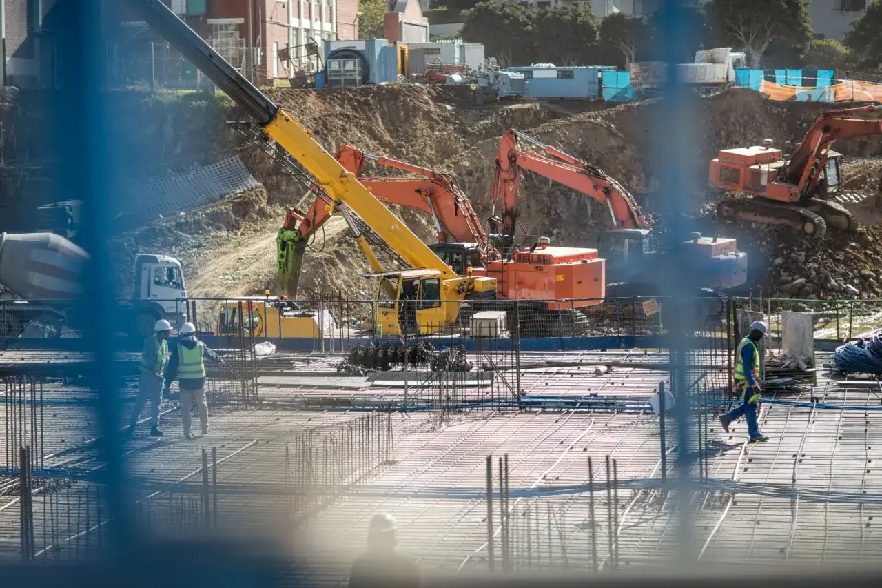 Cantiere con escavatori, gru e camion. Lavoratori supervisionano la preparazione per il getto di cemento, mentre si gestisce lo smaltimento dei rifiuti edili.