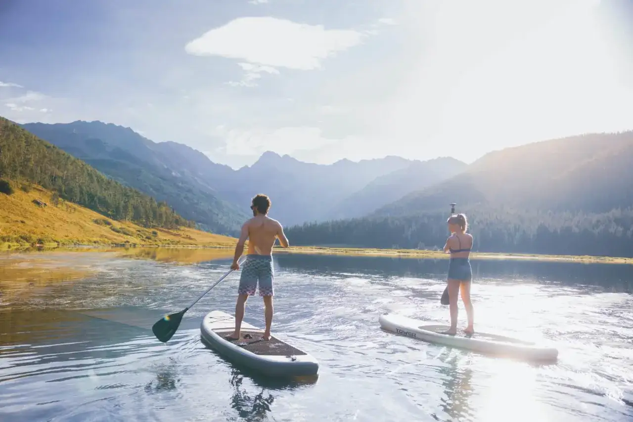 Zwei Personen genießen das Stand-Up-Paddling auf einem ruhigen See vor einer Bergkulisse.