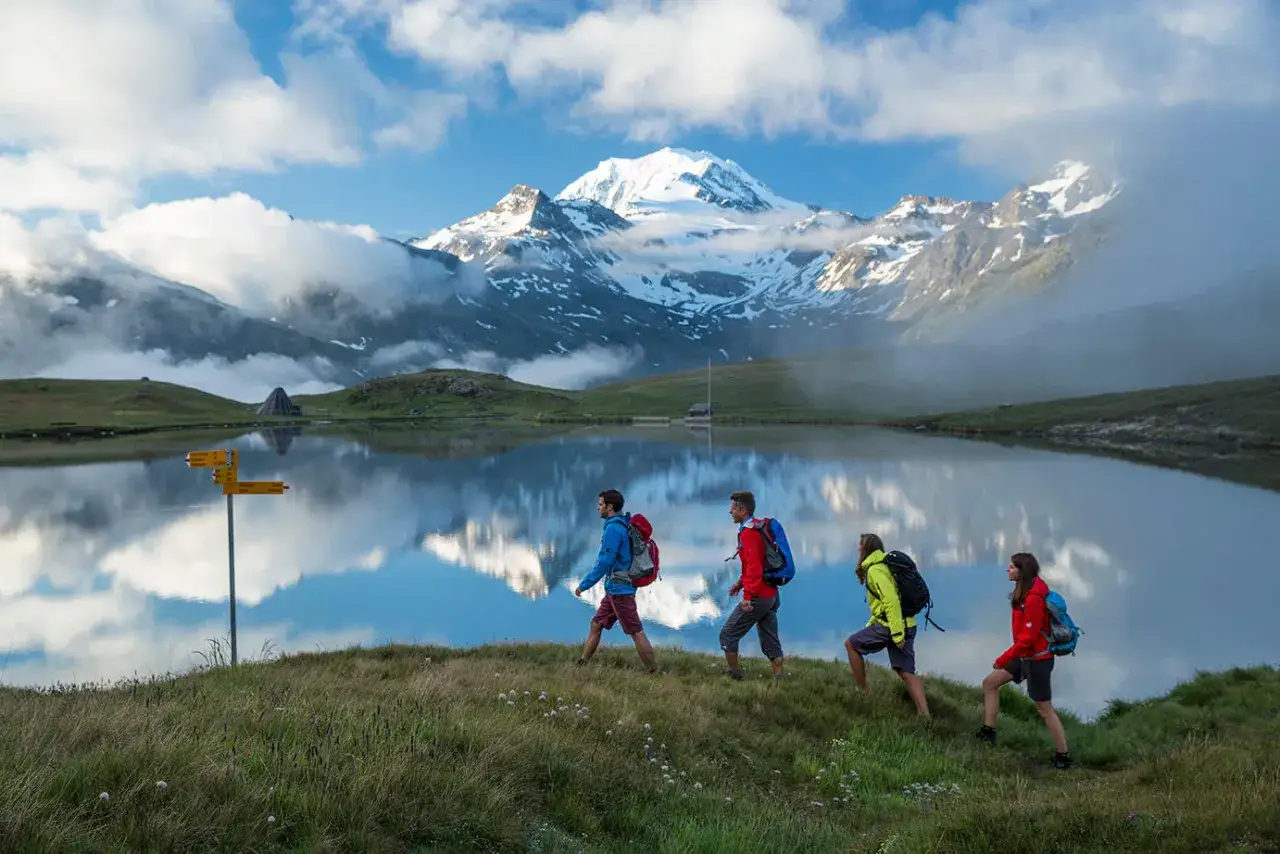 Senderismo en los Alpes suizos. Cuatro excursionistas con mochilas caminan junto a un lago sereno, reflejando montañas nevadas. Voluntariado suiza en su máxima expresión.