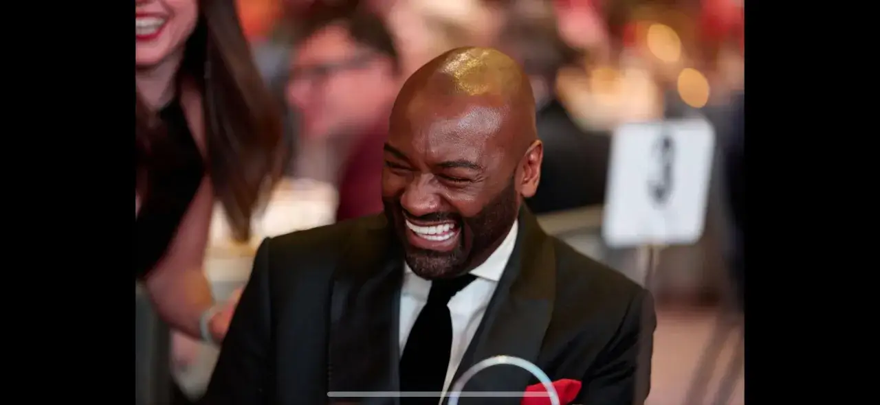 Uomo sorridente in abito elegante, perfetto per fotografare eventi al chiuso. Sfondo sfocato con luci e numero tavolo 3.