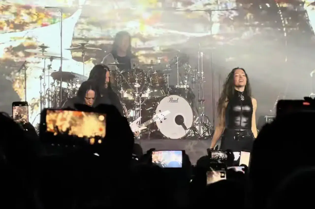 Wokalistka Arch Enemy na scenie, w otoczeniu zespołu, podczas koncertu. Publiczność filmuje występ telefonami.