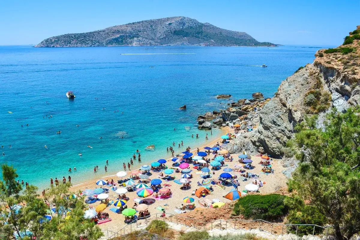 Ein belebter Strand mit vielen bunten Sonnenschirmen und Menschen, die im kristallklaren **Athener Meer** schwimmen. Im Hintergrund eine bergige Insel.