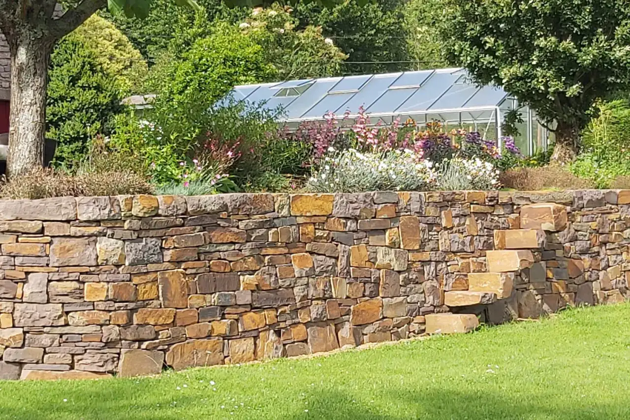 Un beau muret en pierre s&egrave;che soutient un jardin fleuri devant une serre. Des marches en pierre s'int&egrave;grent au muret.