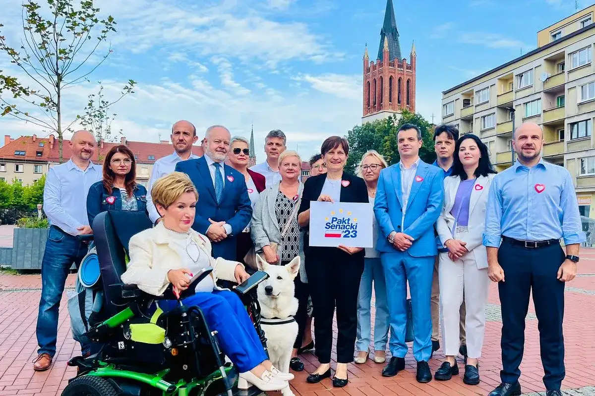 Grupa osób, w tym kandydatka na wózku, pozuje do zdjęcia z hasłem