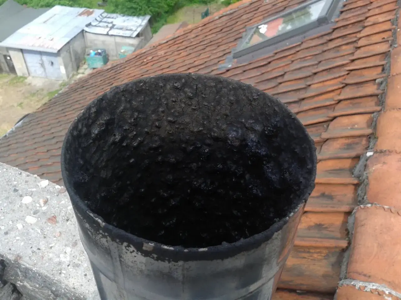 Zatkany komin, czarny osad grozi pożarem i smrodem z komina w pokoju. Dachówki i budynki w tle.