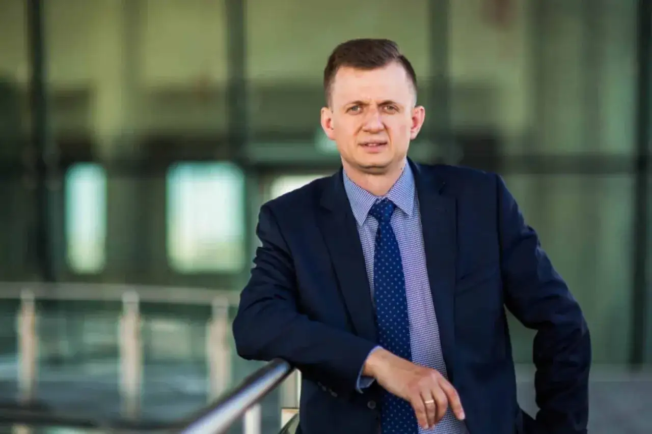 Mężczyzna w garniturze i krawacie opiera się o barierkę na tle nowoczesnego budynku.