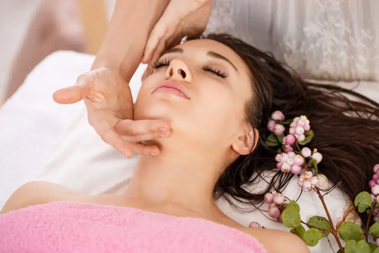 Un massage visage anti rides est prodigué à une jeune femme détendue, ornée de baies roses dans ses cheveux.