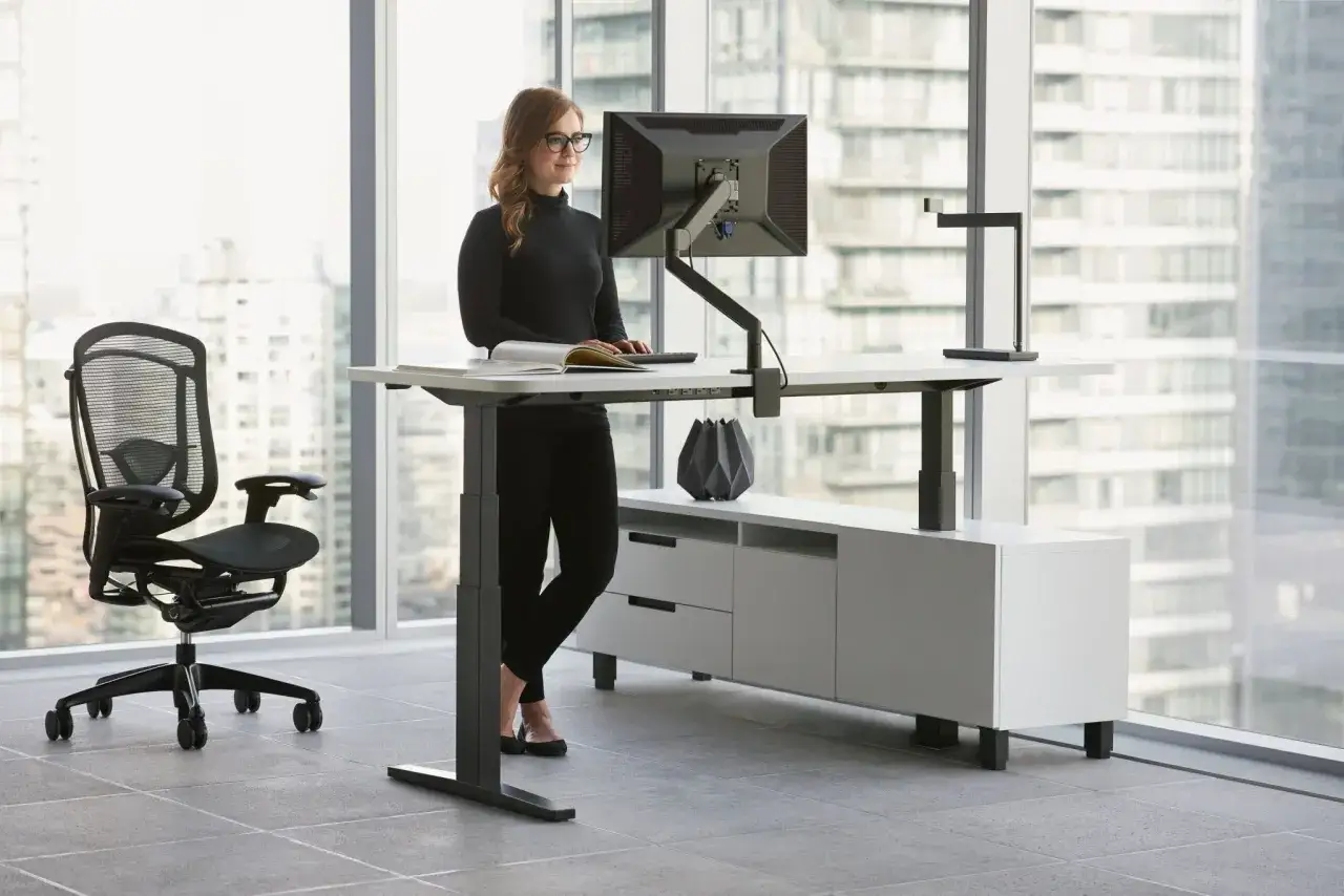 Kobieta pracuje przy biurku z regulacją wysokości, dbając o **ergonomię** pracy. Obok stoi wygodne krzesło biurowe.