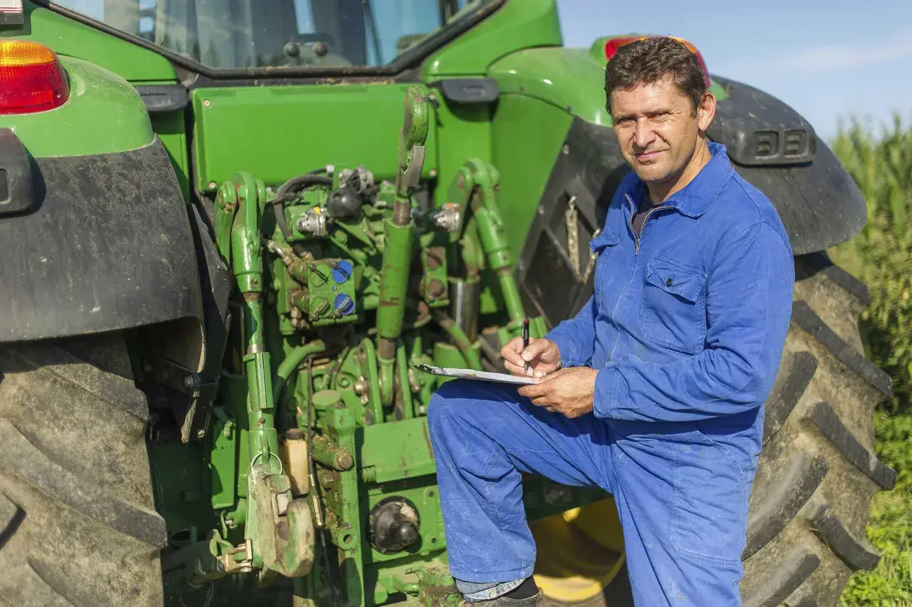 Bauer in blauer Latzhose pr&uuml;ft Aufzeichnungen neben seinem Traktor. Er plant die regelbesteuerung landwirtschaft, um seine Ernte zu optimieren.