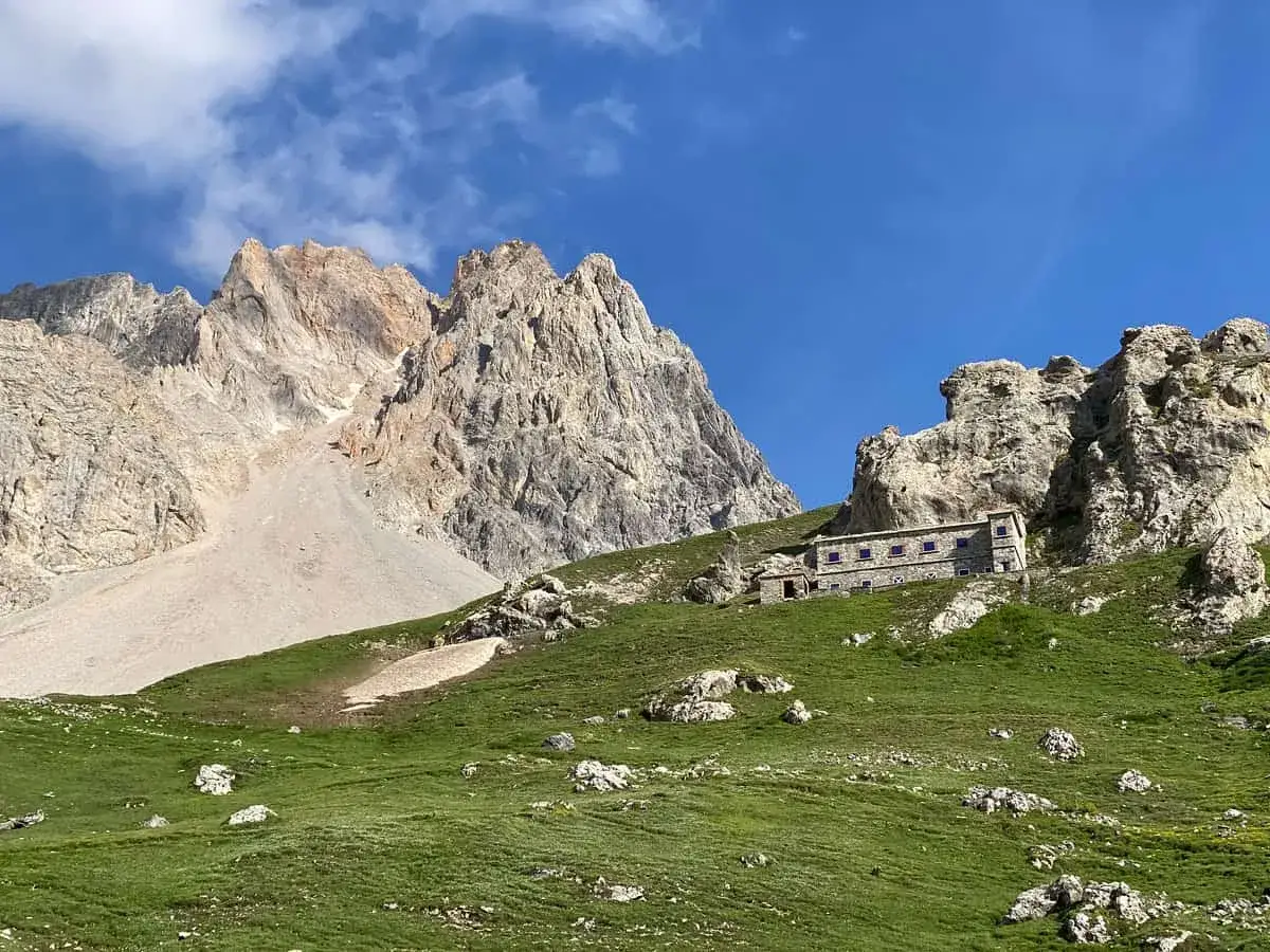 Rifugio alpino in pietra, incastonato nel verde di un prato in **Val di Susa**, dove si trova, con imponenti montagne rocciose sullo sfondo.