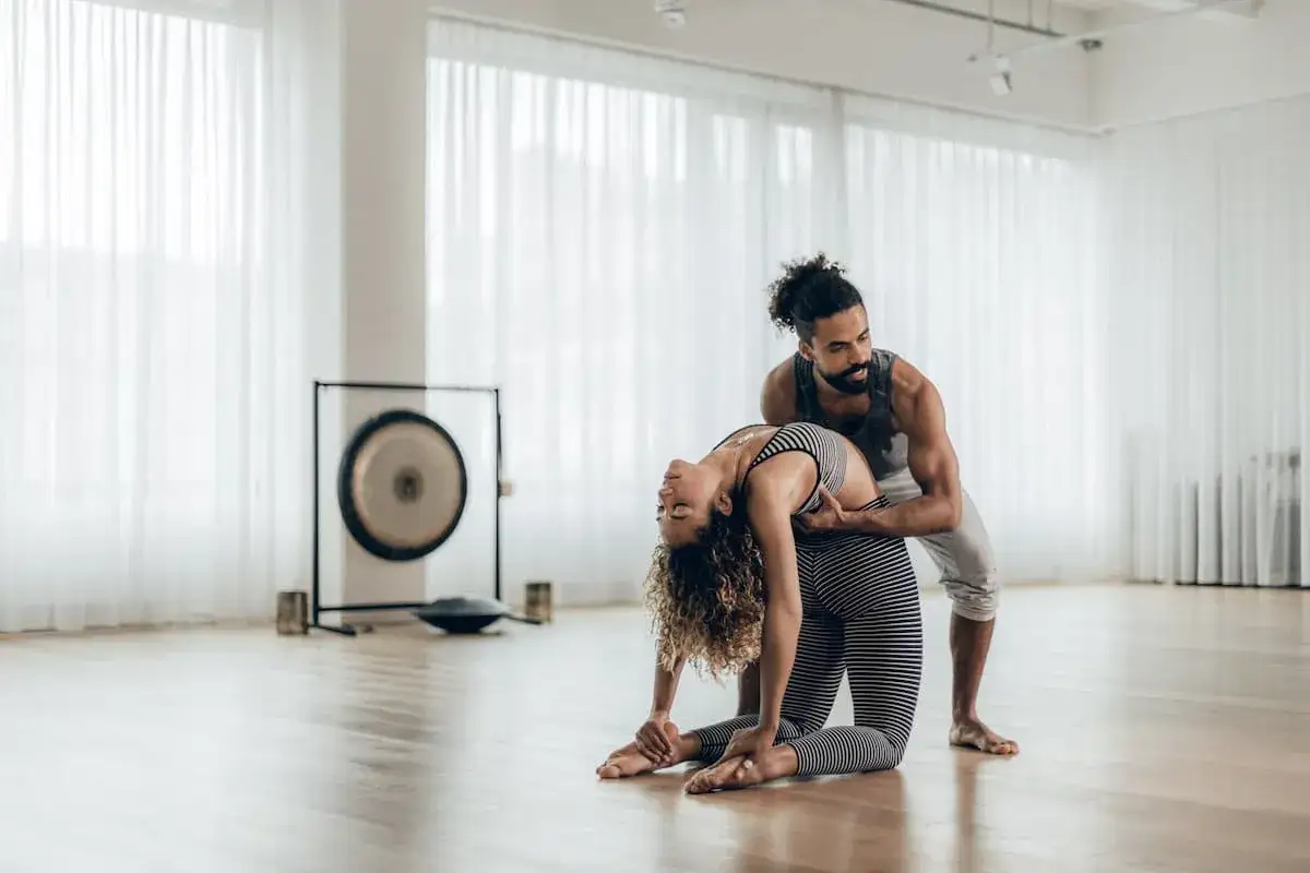 Un maestro de yoga guía a una mujer en una postura de arco profundo en un estudio luminoso.