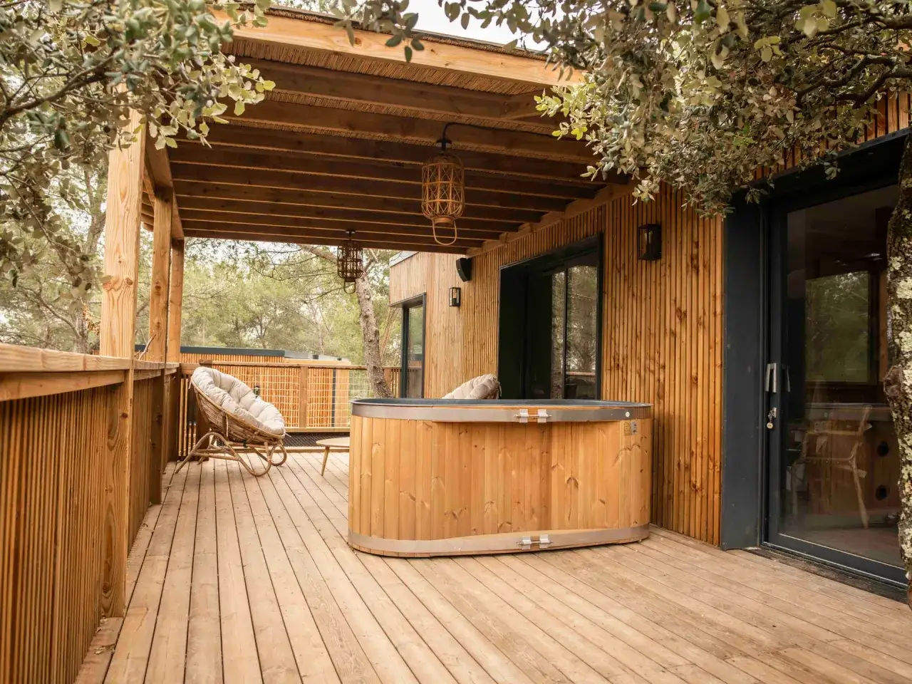 Terrasse en bois d'une cabane var, avec jacuzzi et fauteuil confortable, sous des arbres feuillus.