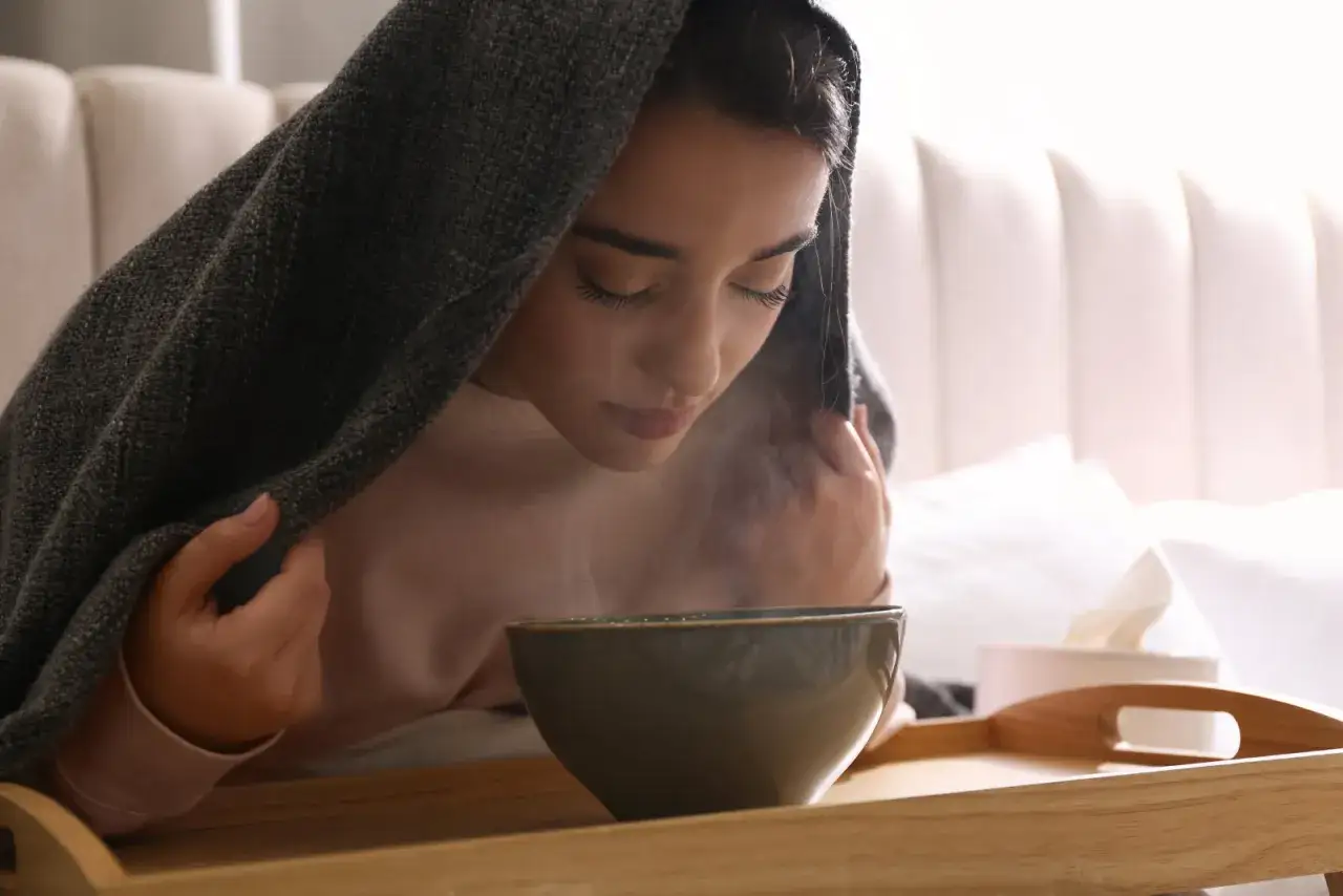 Jeune femme sous une couverture, faisant une inhalation pour son rhume. La vapeur s'échappe d'un bol posé sur un plateau.