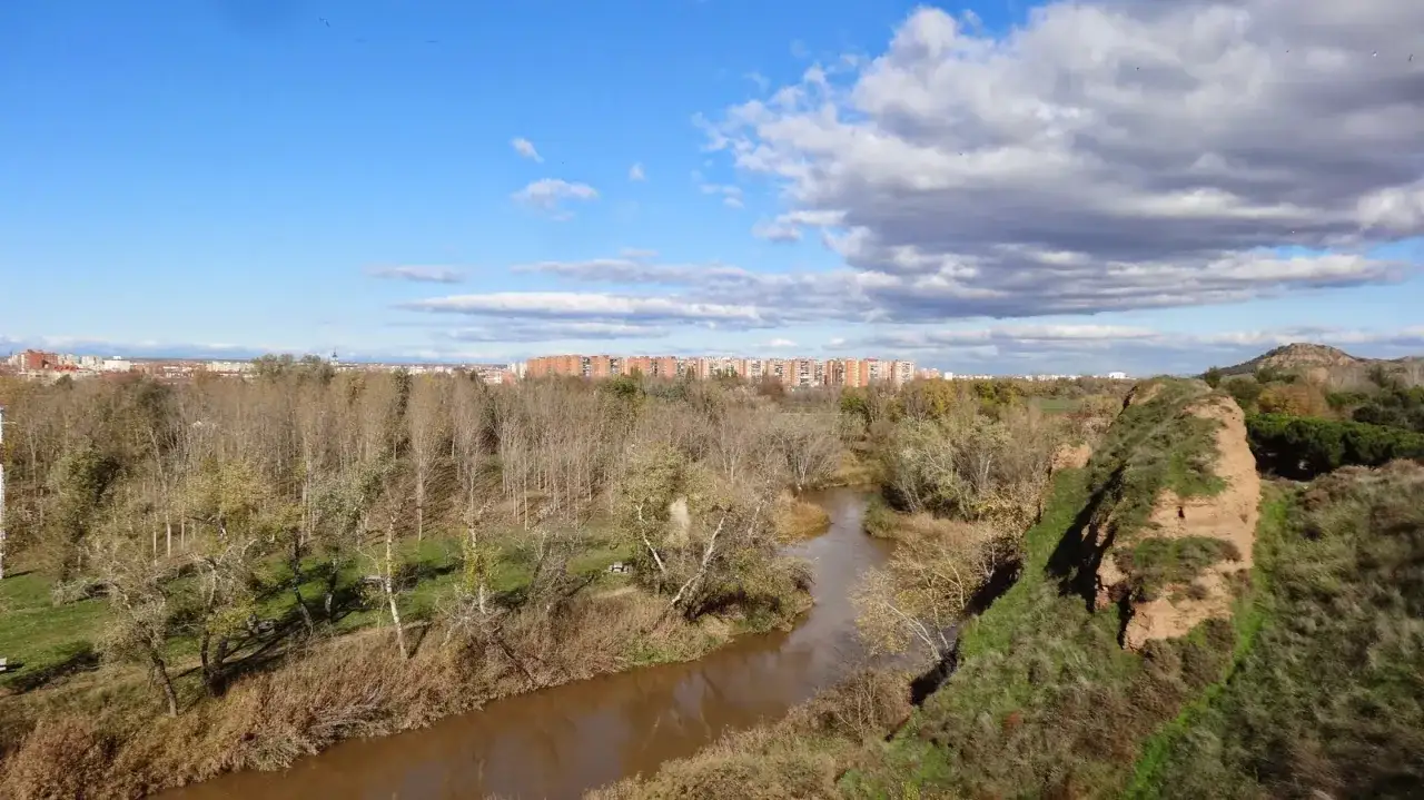 Paisaje otoñal con un río serpenteante y la silueta de edificios urbanos al fondo, con el **cerro del Viso** a la derecha.