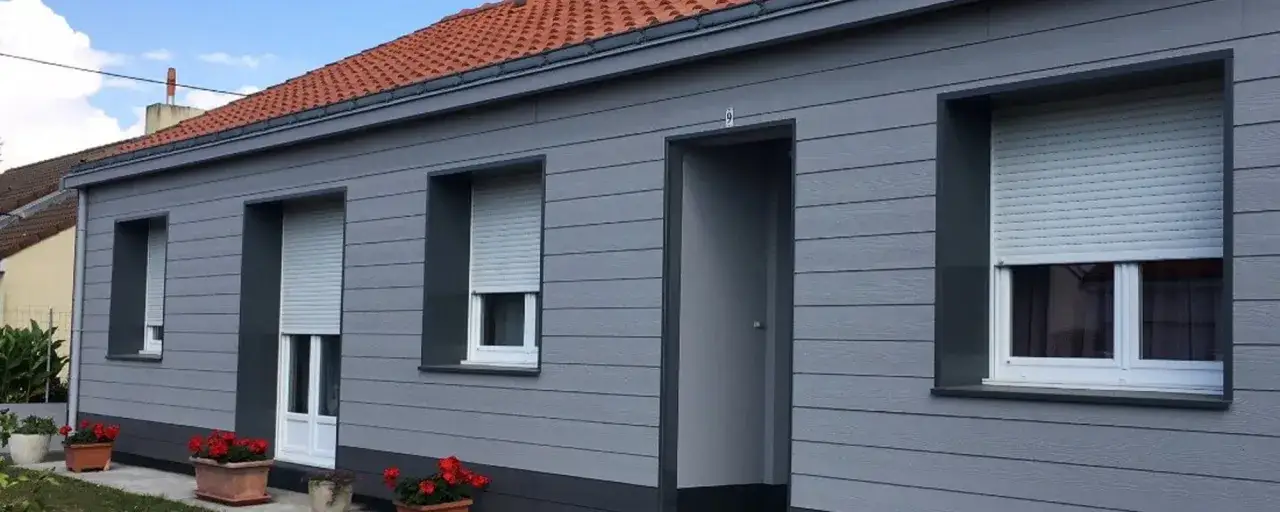 Maison moderne avec bardage gris, volets roulants et jardinières de fleurs rouges. L'isolation par l'extérieur assure confort et esthétique.