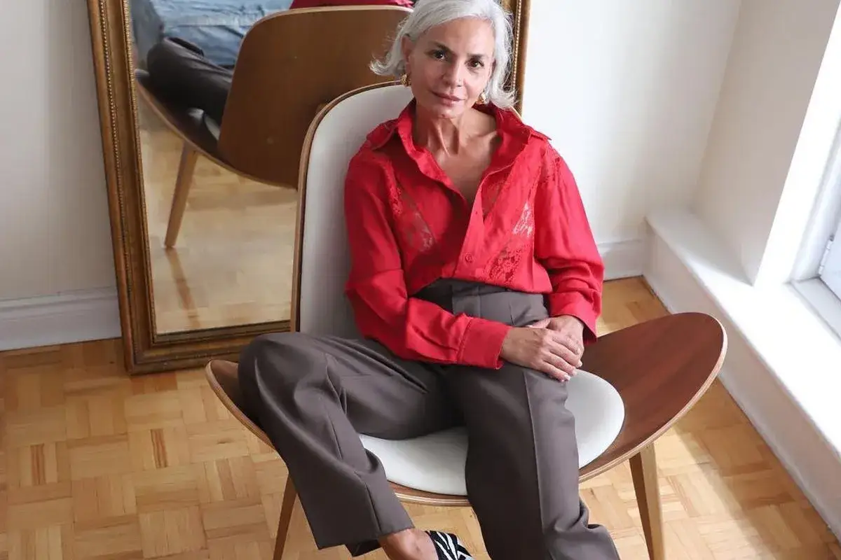 Elegancka kobieta po 50. w czerwonej bluzce i brązowych spodniach, siedzi na krześle. Stylowa moda po 50.