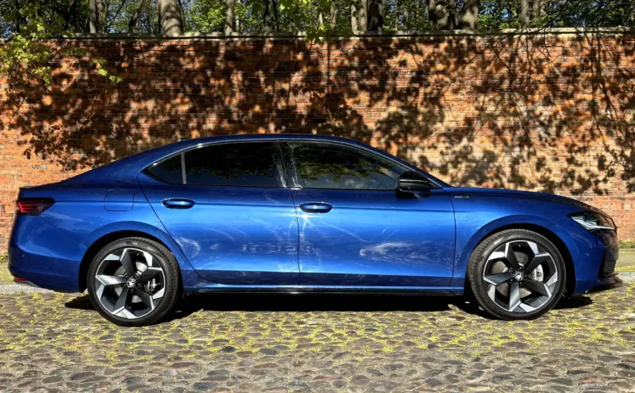 Błękitna, nowa Skoda Superb stoi na brukowanej ulicy. Za nią ceglana ściana z cieniami drzew.