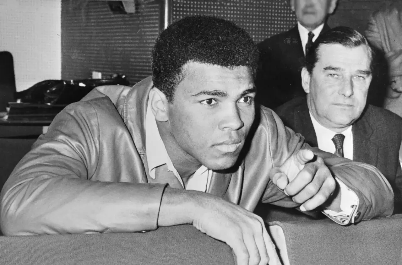 Cassius Clay pekar framåt, redo för nästa utmaning. Bredvid honom sitter en man i kostym.