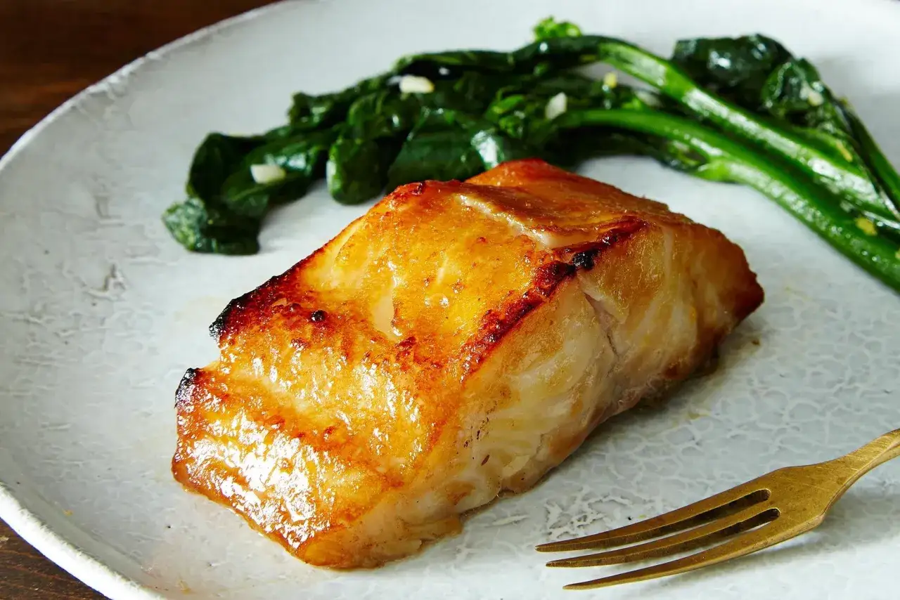 A perfectly seared piece of miso cod recipe, glistening with a golden-brown glaze, served with vibrant green vegetables on a textured plate.