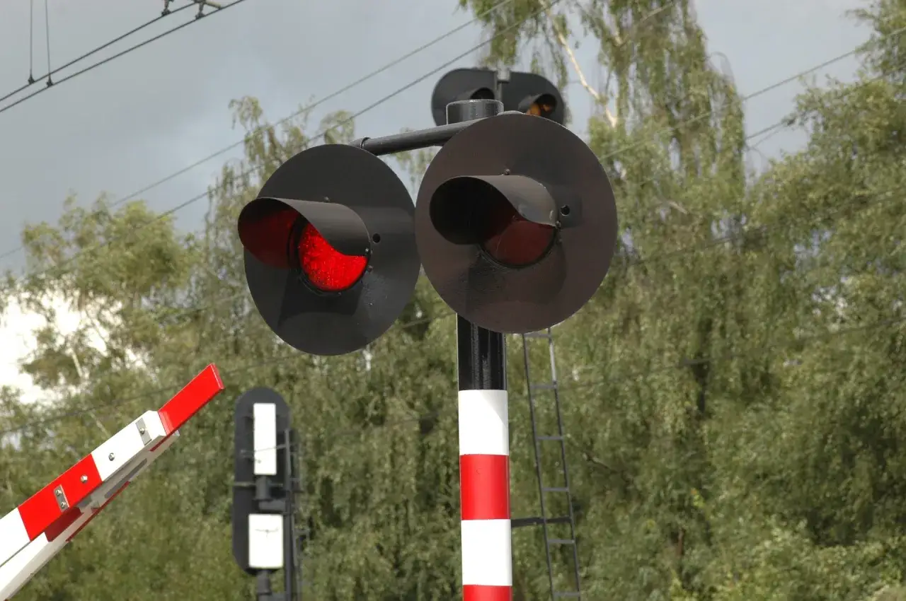 Czerwone światła na przejeździe kolejowym ostrzegają przed nadjeżdżającym pociągiem. Bariera jest opuszczona, a drzewa tworzą zielone tło.