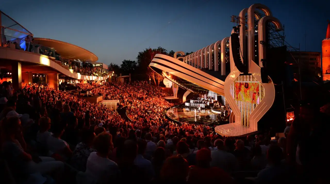 Koncert w amfiteatrze w Polsce. Tłum widzów siedzi na widowni, podziwiając nowoczesną scenę oświetloną wieczorem.