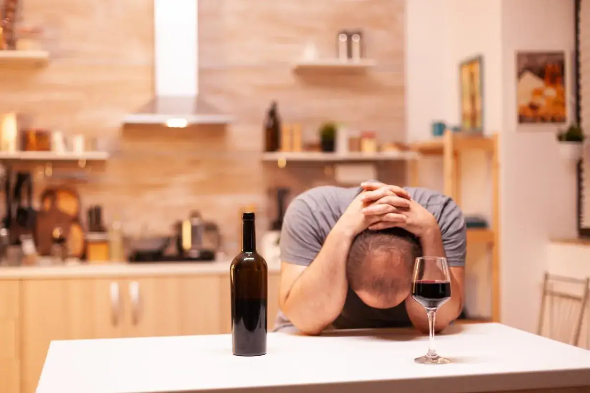 Zrozumienie, jak rozmawiać z alkoholikiem aby przestał pić, jest kluczowe. Mężczyzna z głową w dłoniach przy butelce wina symbolizuje walkę z nałogiem.