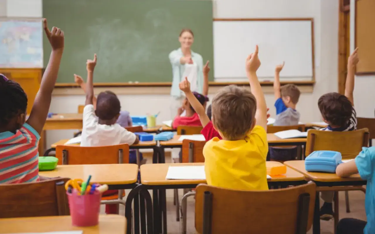 W szkole podstawowej uczniowie z radością podnoszą ręce, chcąc odpowiedzieć na pytanie nauczycielki.