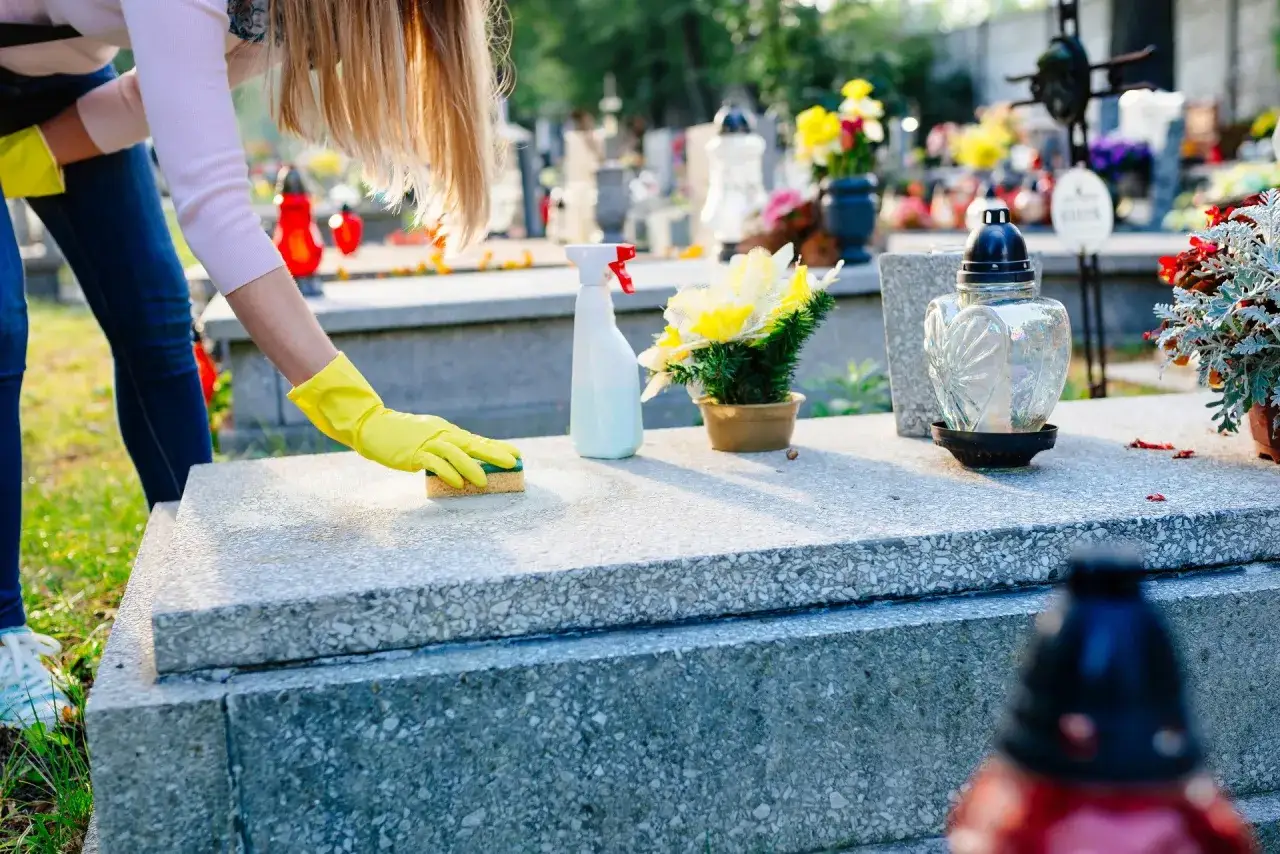 Kobieta w żółtych rękawiczkach czyści pomnik gąbką. Na nagrobku stoi wazon z kwiatami i znicz. Pytanie: czym zmyć wosk z pomnika?