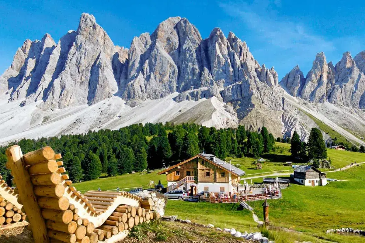 Un rifugio alpino e altri piccoli paesi nel Val di Funes, con imponenti montagne sullo sfondo e un prato verde.