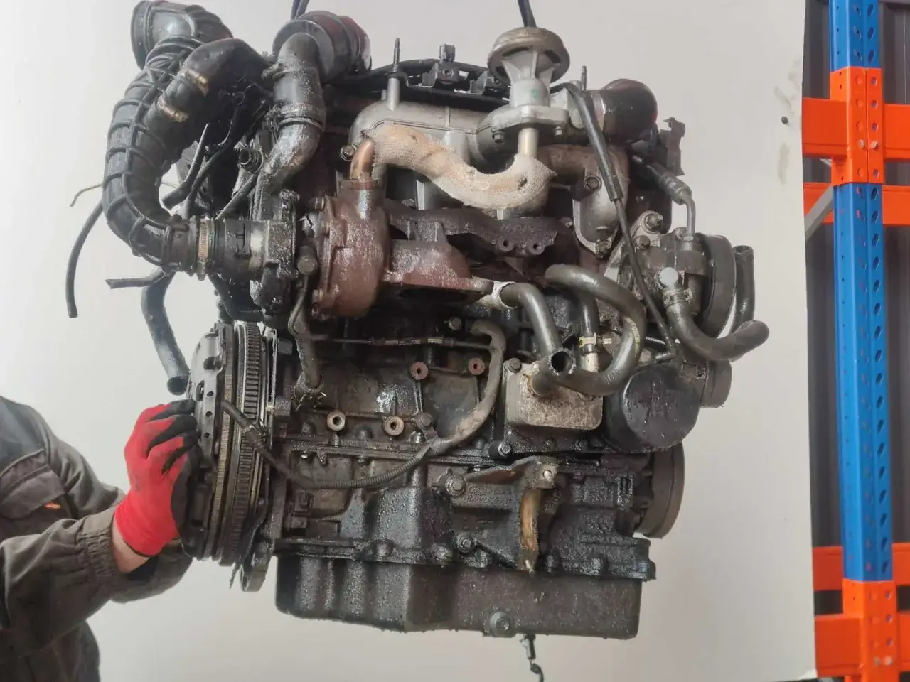 Silnik 1.8 TDCI, brudny, z turbosprężarką, w trakcie demontażu. Ręka w czerwonej rękawicy przy sprzęgle.