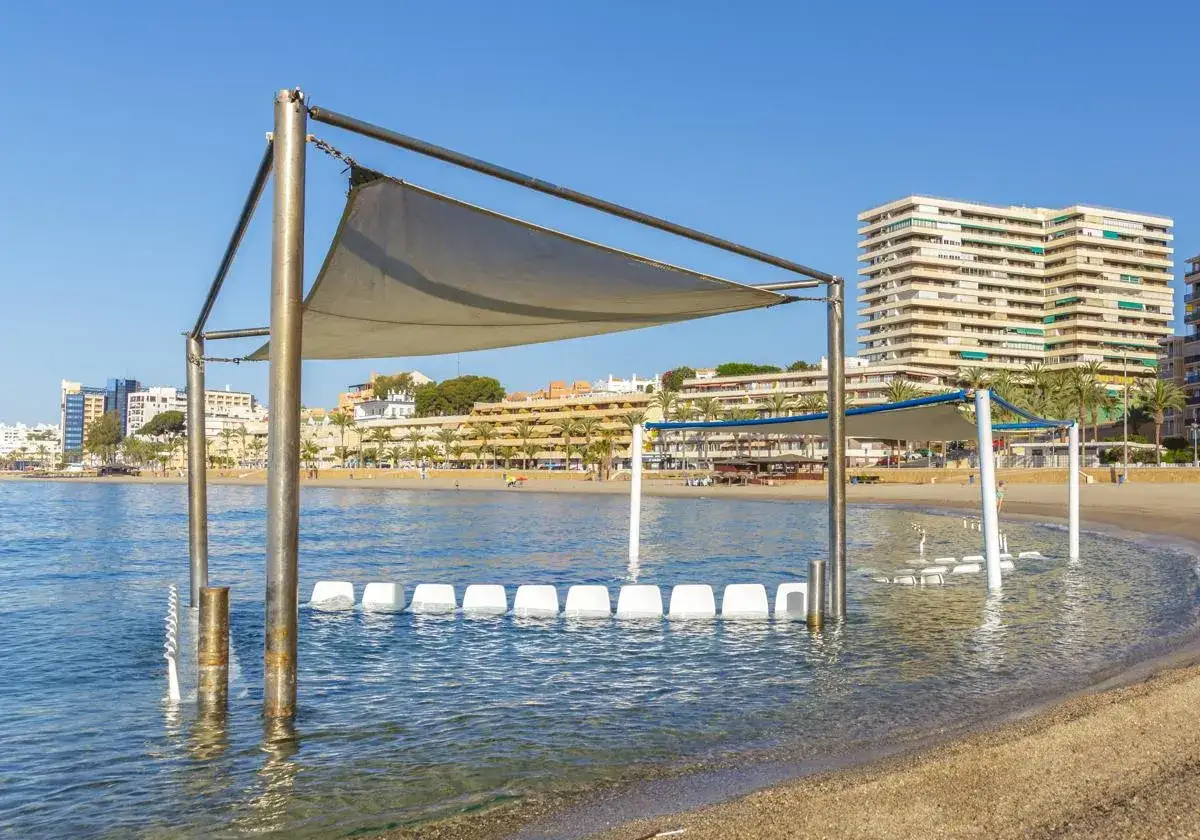 Sillas blancas sumergidas en el mar en el puerto de Roquetas de Mar, con edificios y palmeras al fondo bajo un cielo azul.