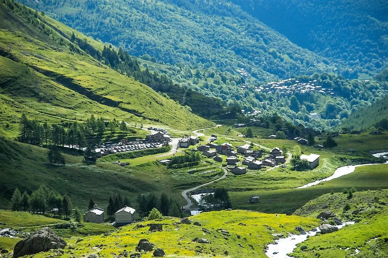 Un villaggio alpino nel **Pian della Regina**, con case in pietra e un torrente che scorre tra prati verdi e boschi lussureggianti.