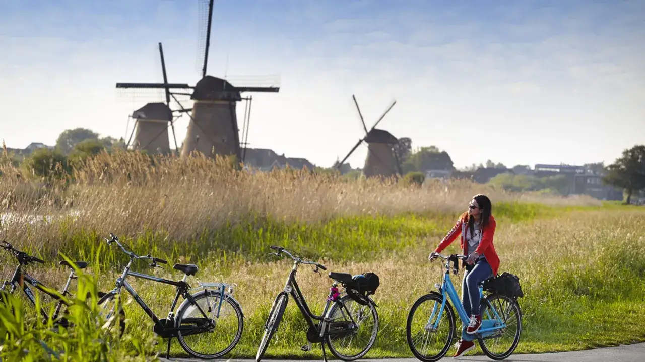 Mujer pedalea en bici por un campo con molinos de viento al fondo, una escena típica de Holanda en bici.