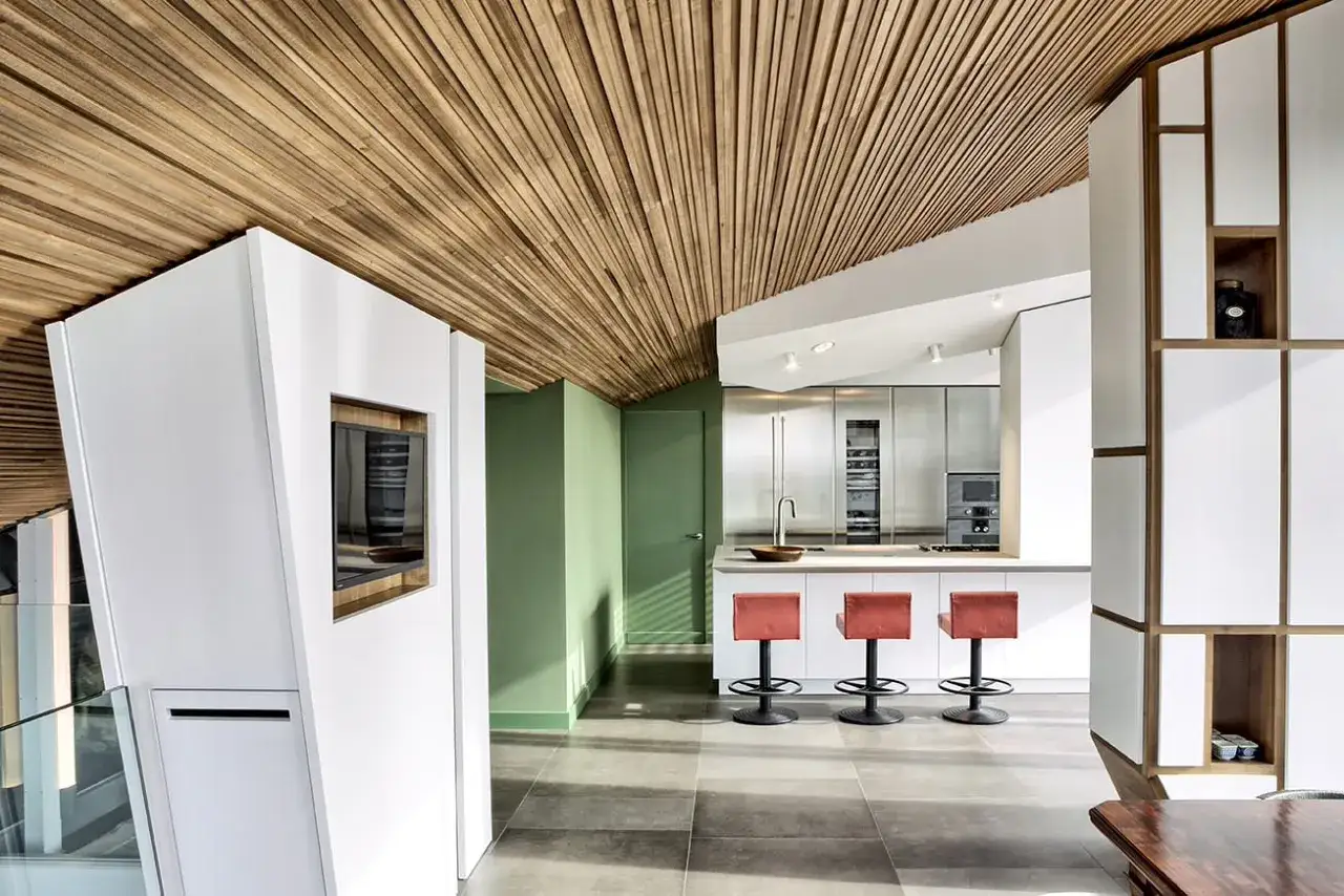 Moderne K&uuml;che mit Holzbalken an der Decke, die verkleidet ist. Wei&szlig;e Elemente und rote Barhocker.