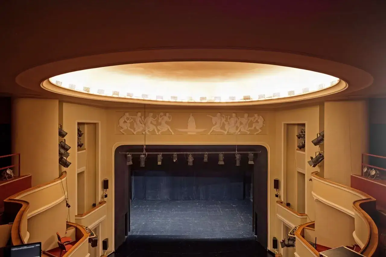 Wnętrze teatru w Kaliszu. Scena z kurtyną, balkony, reflektory i dekoracja nad sceną. Sprawdź repertuar!
