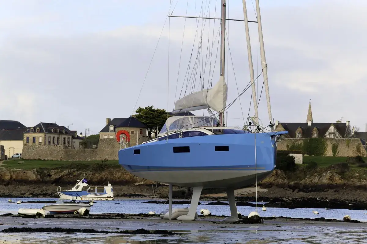 Un voilier bleu futuriste, un dériveur intégral, repose sur ses béquilles sur une plage bretonne. Au loin, des maisons et une église.
