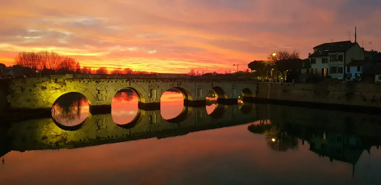 Ponte di Tiberio a Rimini al tramonto, con riflessi sull'acqua e un cielo infuocato. Lo skyline di Rimini &egrave; incantevole.
