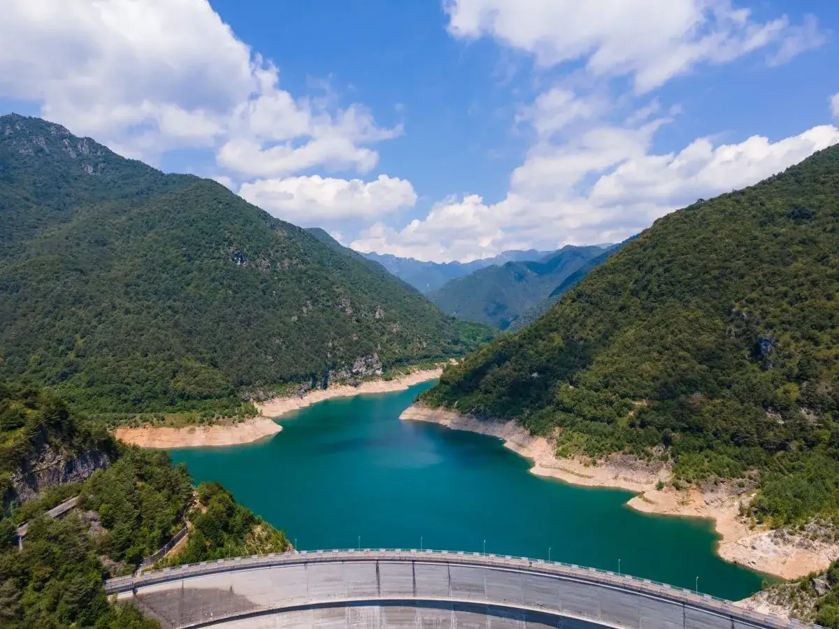 Imponente diga sul lago di Valvestino, circondato da montagne verdi e un cielo azzurro con nuvole bianche.