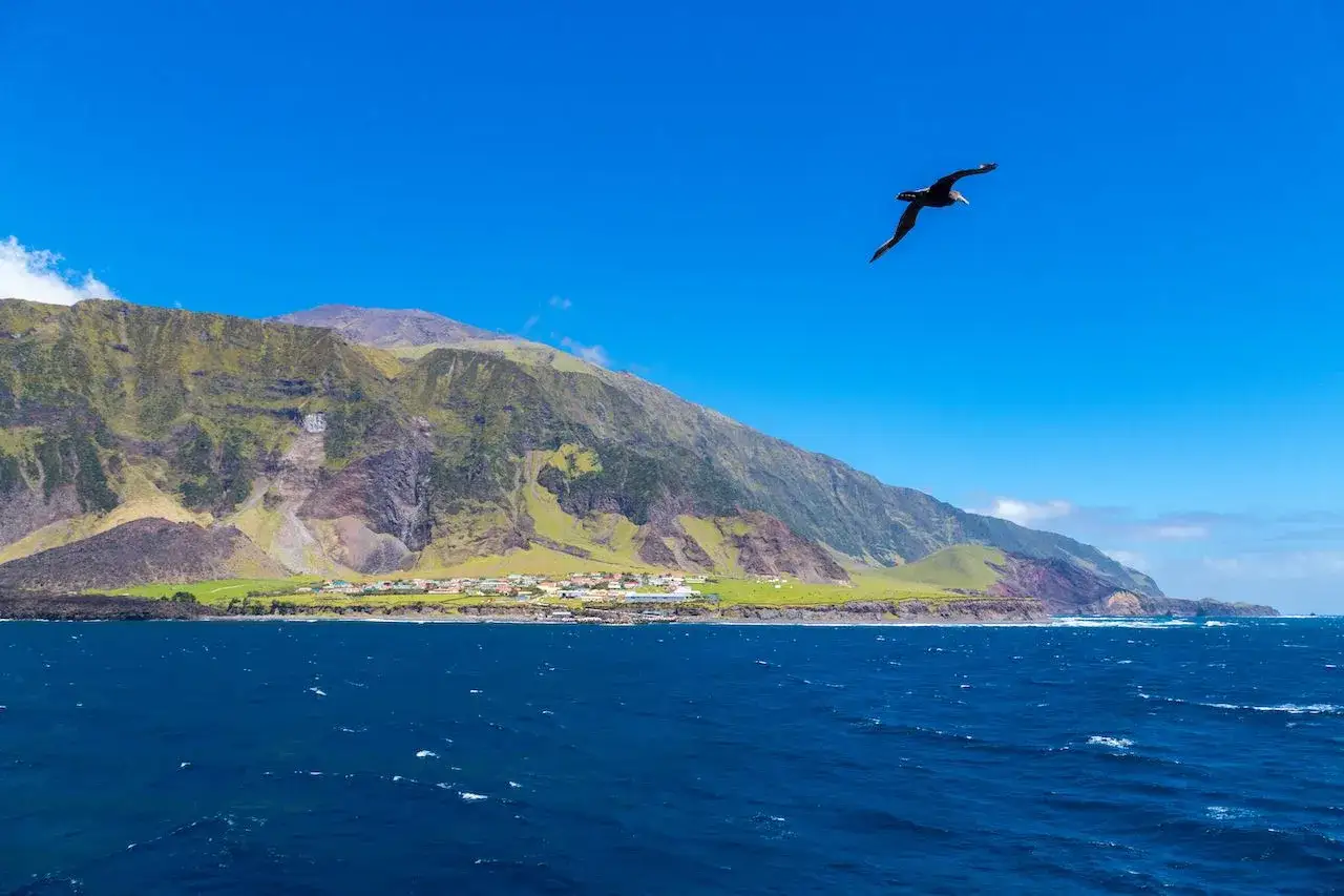 Un ave surca el cielo azul sobre el asentamiento de Tristan de Acuña, con sus casas coloridas a los pies de montañas verdes y escarpadas.