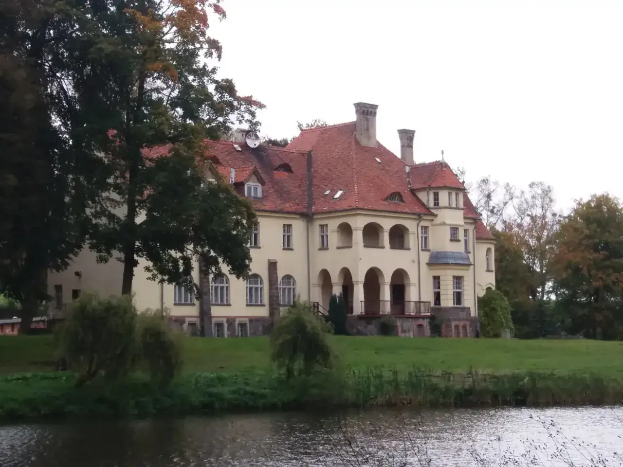 Ż&oacute;łty pałac z czerwonym dachem, otoczony zielenią, stoi nad spokojną wodą.