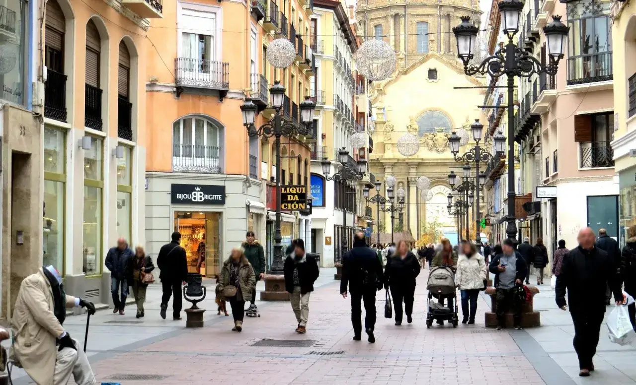 Gente pasea por una calle comercial con edificios históricos y un gran templo al fondo, evocando el encanto de Zaragoza.