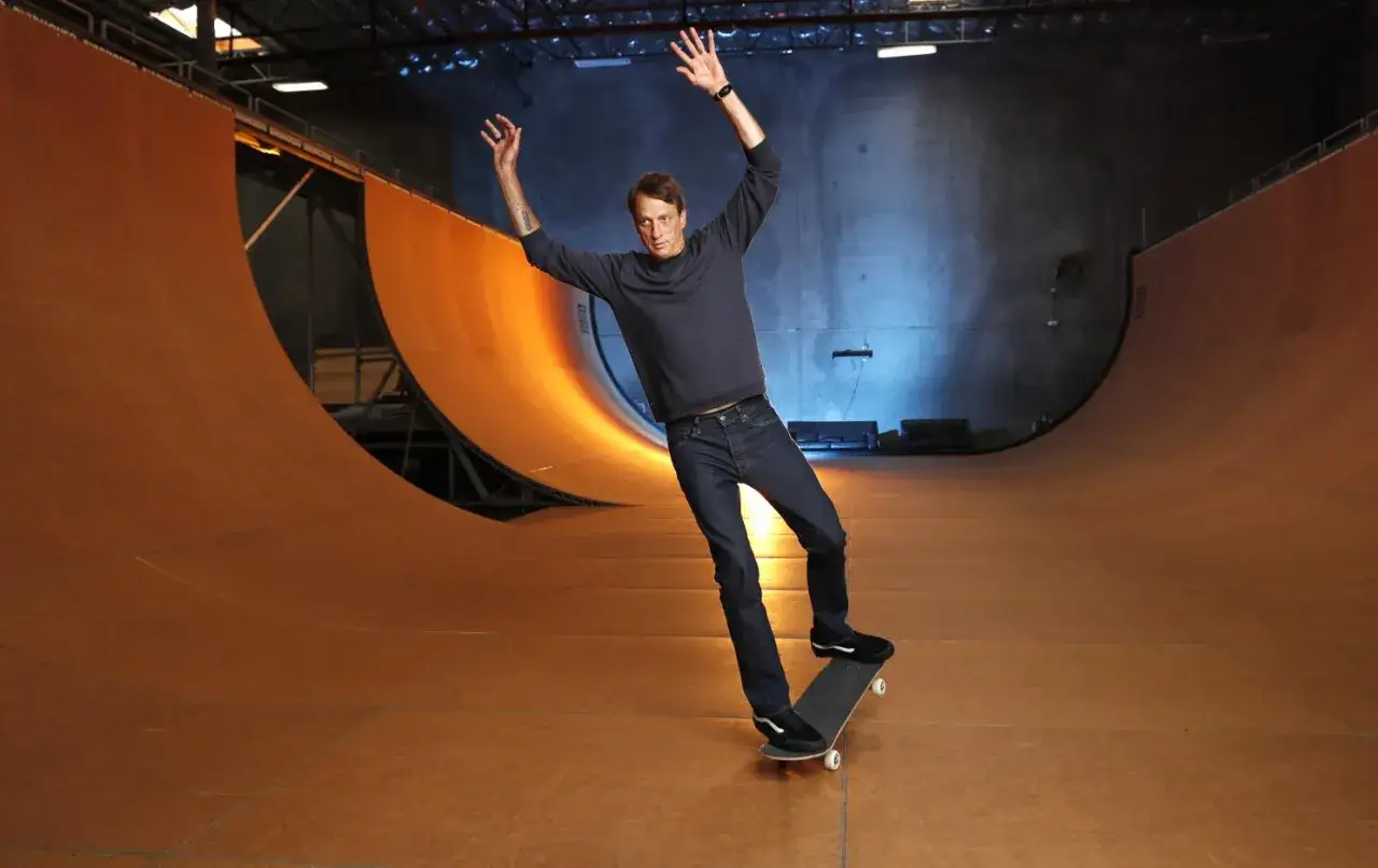 Tony Hawk w swoim filmie na deskorolce, balansuje na krawędzi rampy w hali.