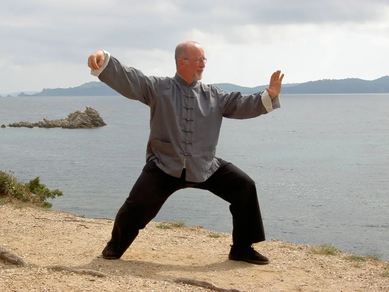 Un homme pratique la gym chinoise pour seniors au bord de la mer, dans une posture de Tai Chi.