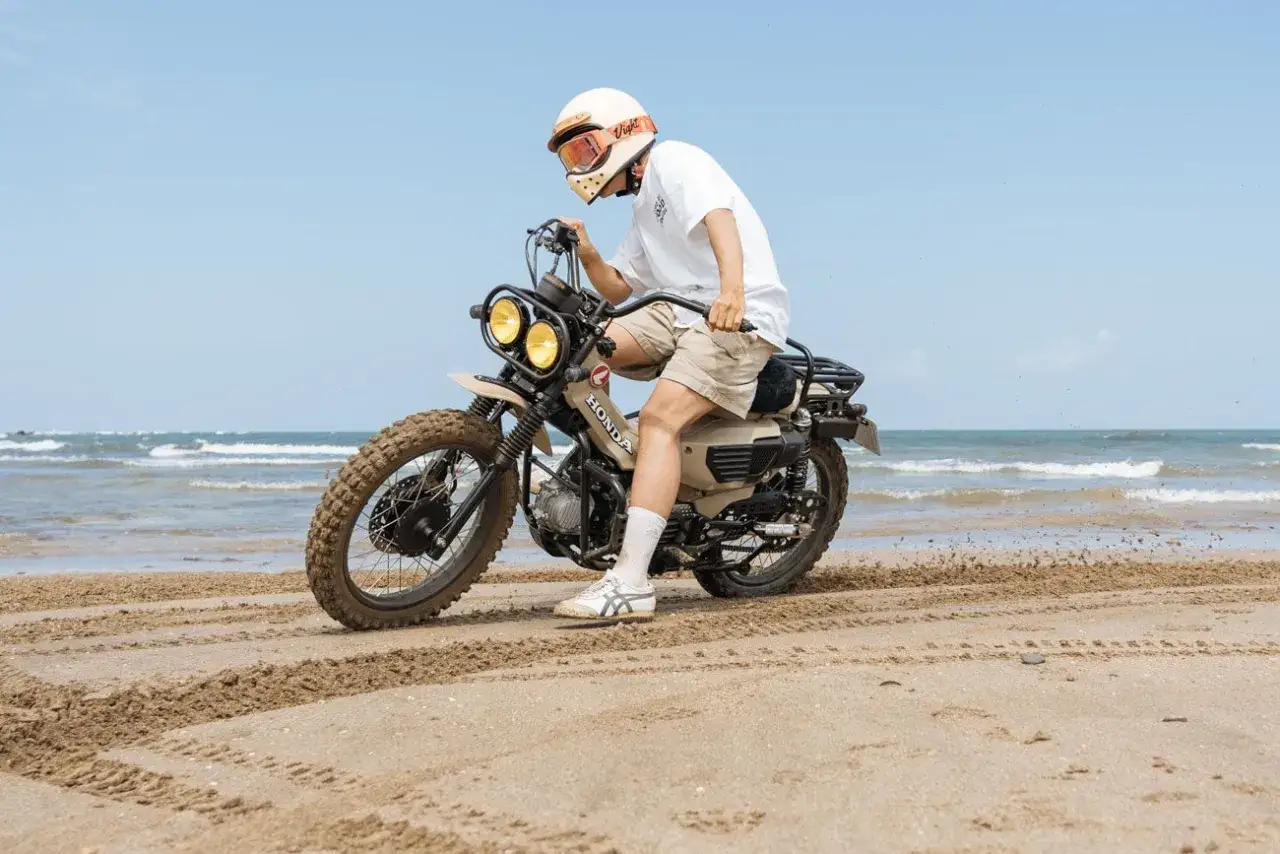Mężczyzna w kasku i okularach jedzie piaskową Honda CT 125 po plaży. W tle widać morze i błękitne niebo.
